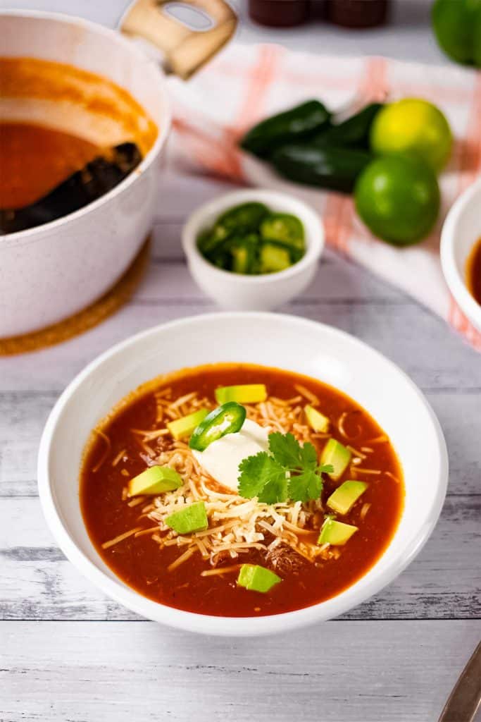 Taco soup in a white soup bowl on a table with other ingredients, jalapeño peppers and limes.