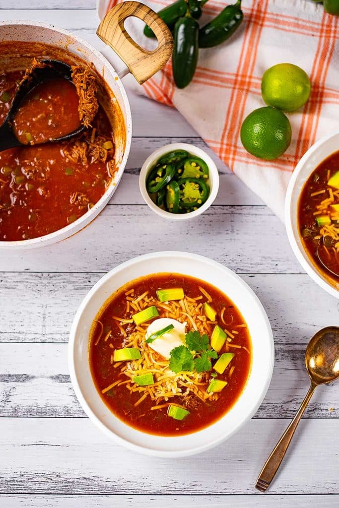 Keto taco soup in a white bowl on a table with limes, jalapenos, puled pork with a bronze spoon to the side.