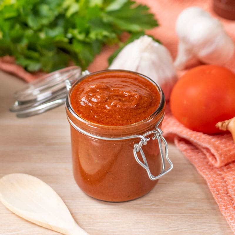 Keto Enchilada Sauce in a glass swing top jar on a wooden table surround by whole ingredients
