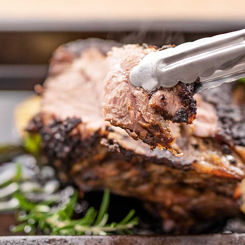 Image show a cooked lamb shoulder in a roasting pan with fresh rosemary, and mint.. One slice is held up to the camera.