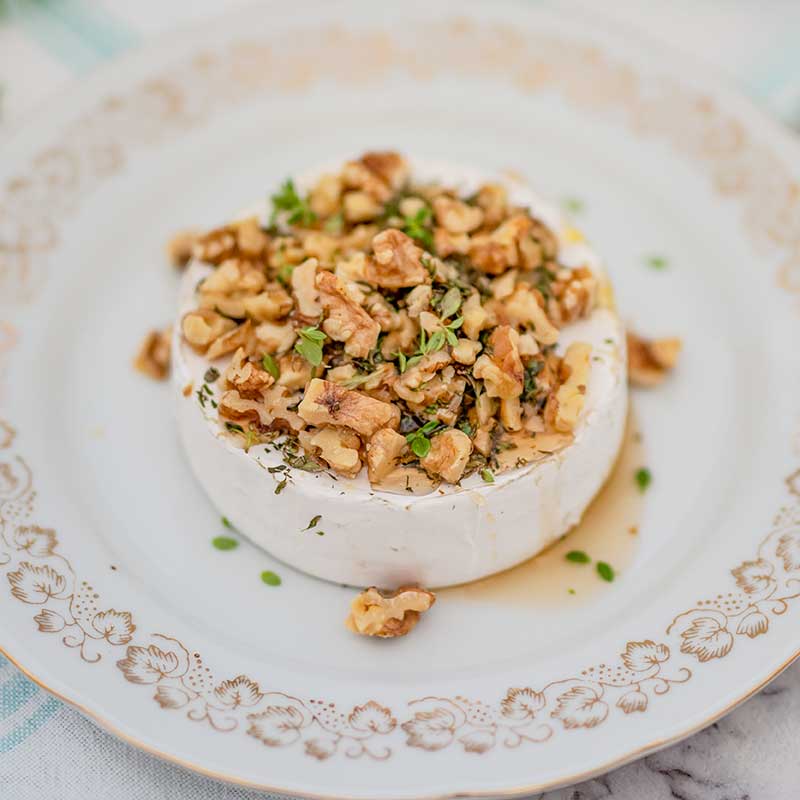 Image of a wheel of brie cheese on a white plate with gold trim. The brie is topped with walnuts, thyme and sugar free maple syrup