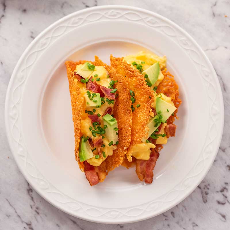 Imago shows two keto breakfast tacos on a white plate. The tacos are filled with bacon, scrambled eggs, avocado and chives.