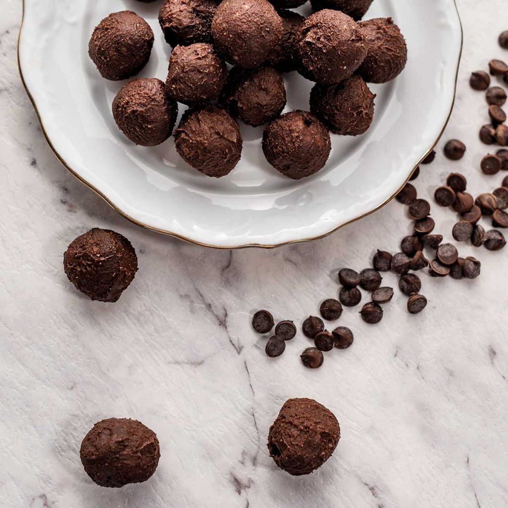 Image shows keto brownie fat bombs on a white marble bench with sugar free chocolate chips sprinkled around and some on a white plate