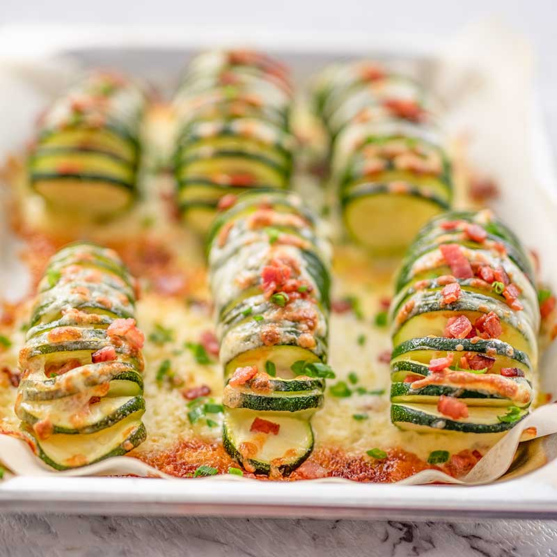 Image shows 6 pieces of keto hasselback zucchini stuffed with bacon and cheese sitting on a cookie sheet