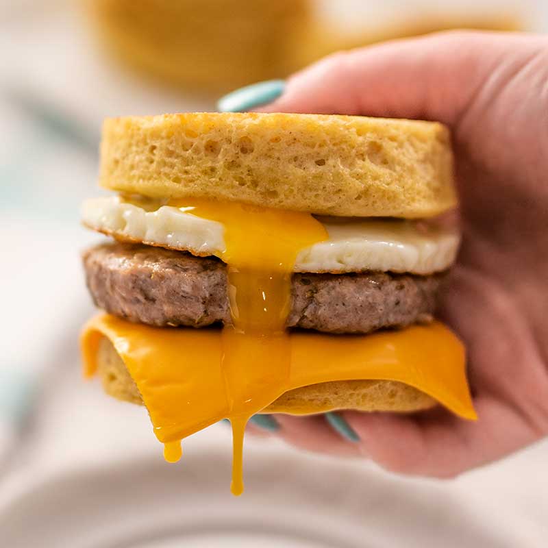 Image shows a keto sausage and egg muffin being held up to the camera, there is runny egg yolk dripping down the front