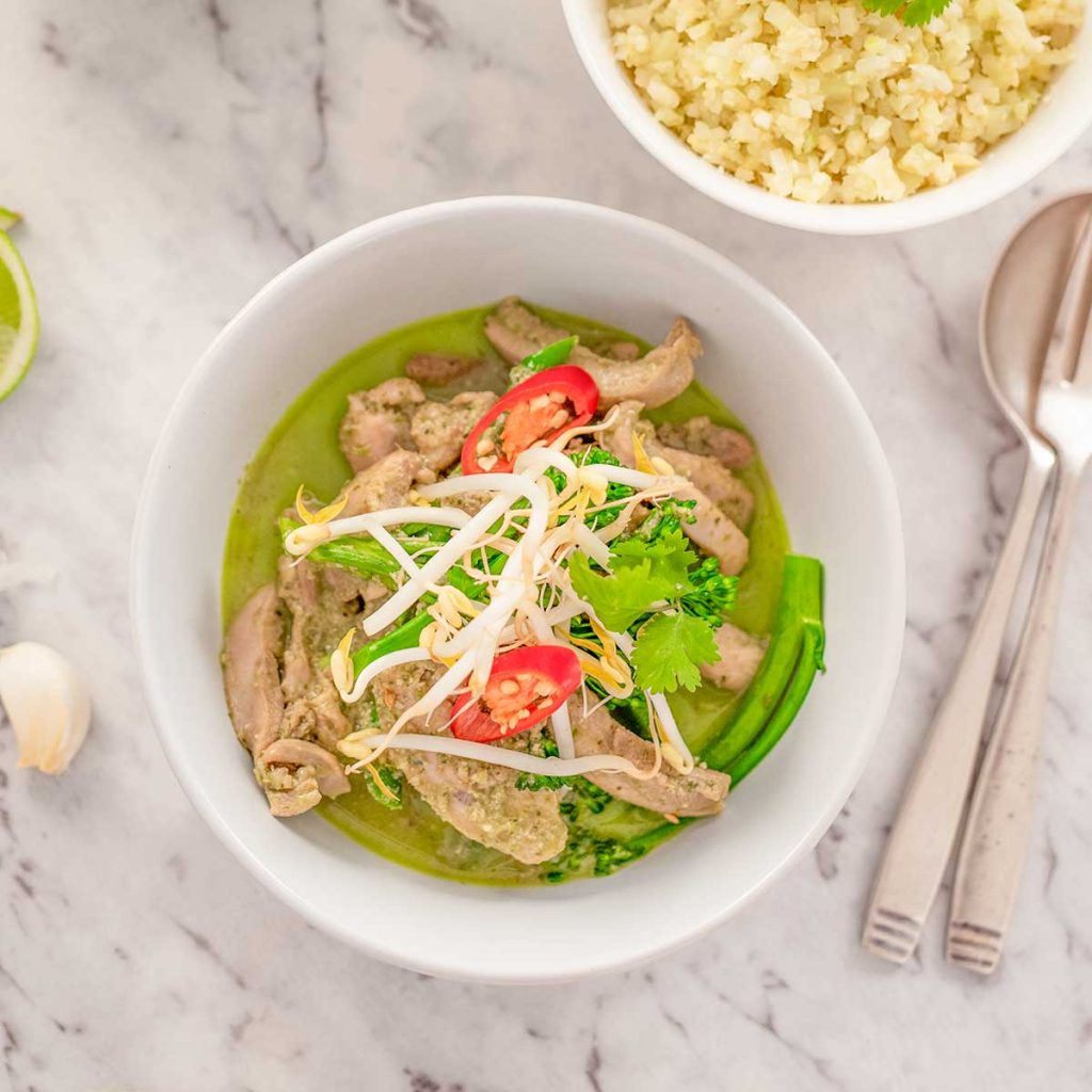 Image shows keto thai chicken green curry in a white bowl topped with bean sprouts, cilantro and a slice of red pepper. There are utensil and a bowl of cauliflower rice in the background