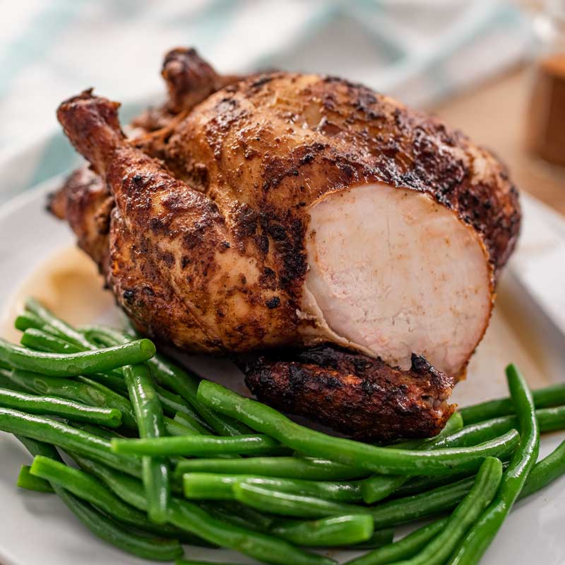 Image of a rotisserie chicken with a slice removed on a white plate with green beans around it on one side and a carving knife on the other.