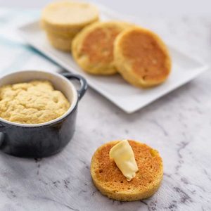 Image shows a buttered keto english muffin, another is behind it in a ramekin and a plate full in the background