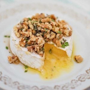 Image of a wheel of brie cheese on a white plate topped with walnuts, thyme and sugar free maple syrup with one slice remove. The melted cheese is oozing out.