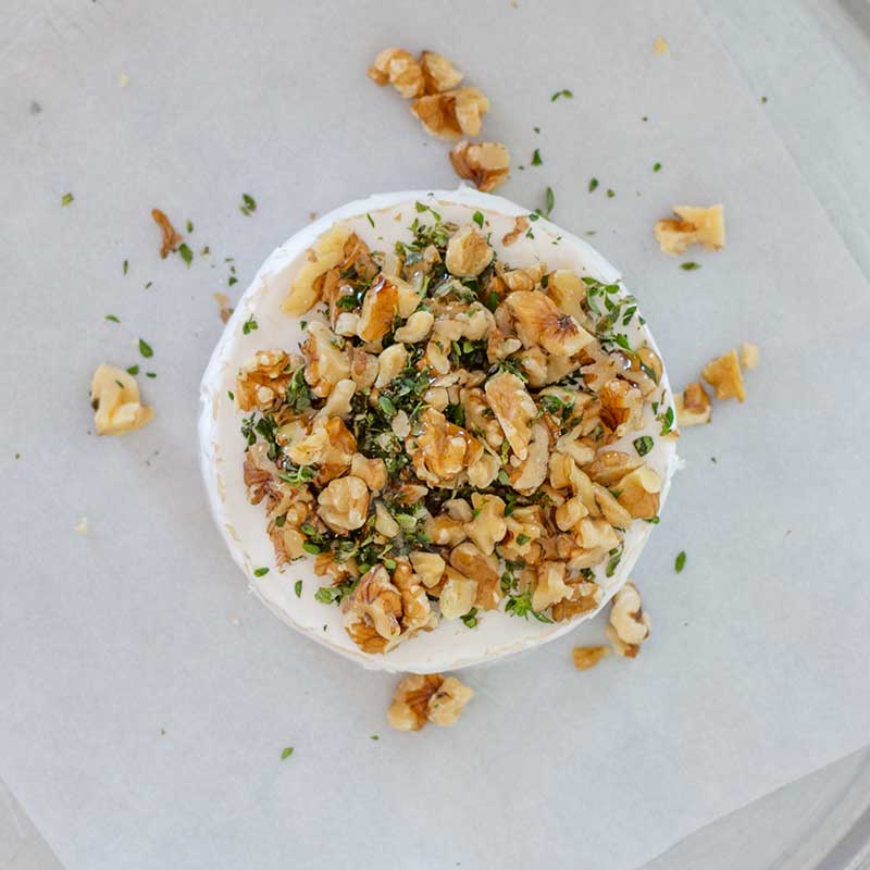 Image of a wheel of brie on a lined baking pan topped with walnuts, thyme and sugar free maple syrup