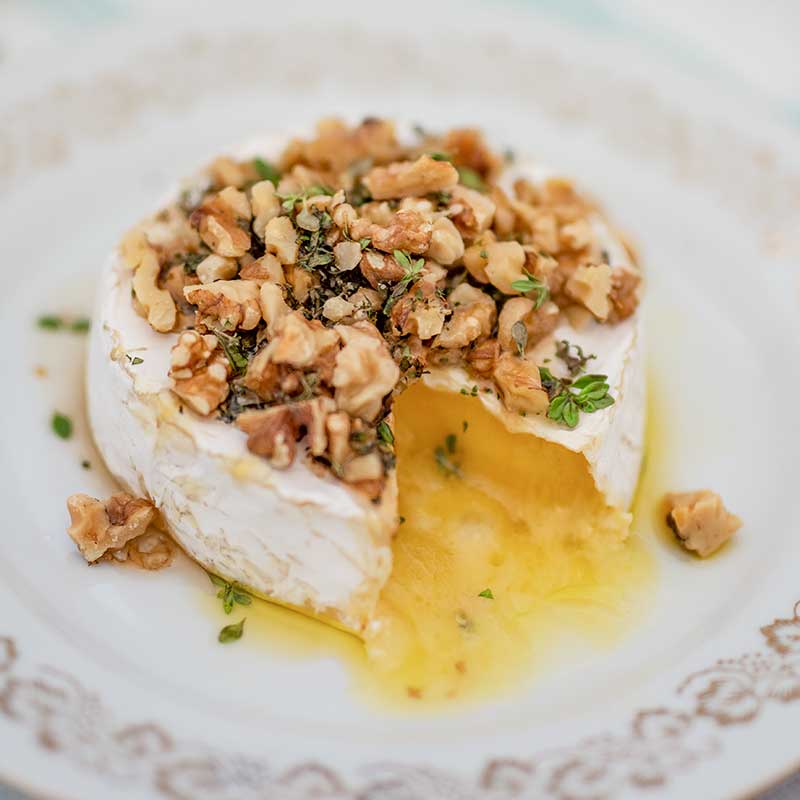 Image of a wheel of brie cheese on a white plate topped with walnuts, thyme and sugar free maple syrup with one slice remove. The melted cheese is oozing out.