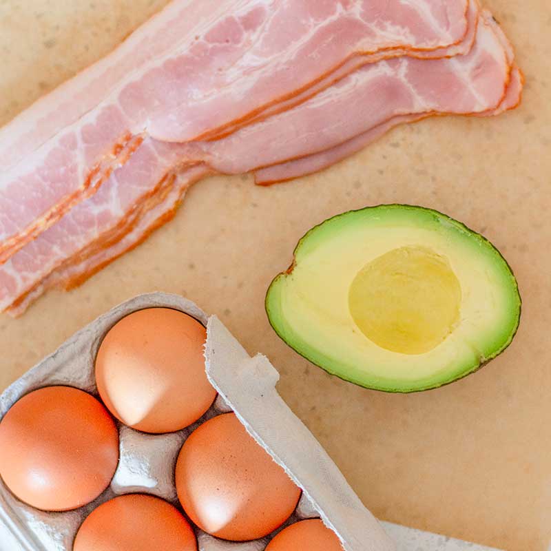 Image of bacon, eggs and half an avocado on a light brown background