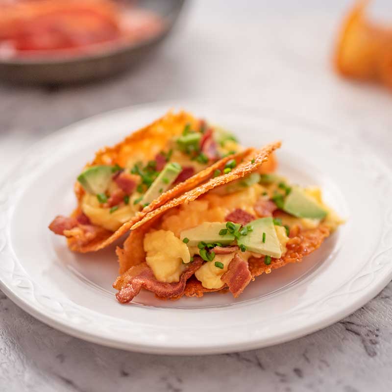 Two keto breakfast tacos on a white plate. The tacos are filled with bacon, scrambled eggs, avocado and chives.