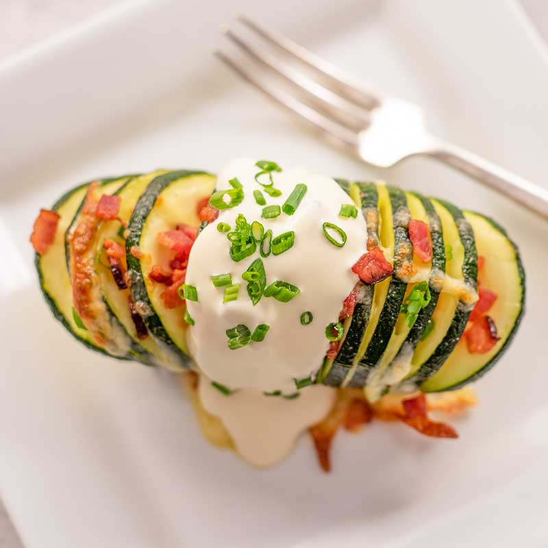 Image of a keto hasselback zucchini stuffed with cheese and bacon with sour cream and chives on top, sitting on a white plate with a silver fork