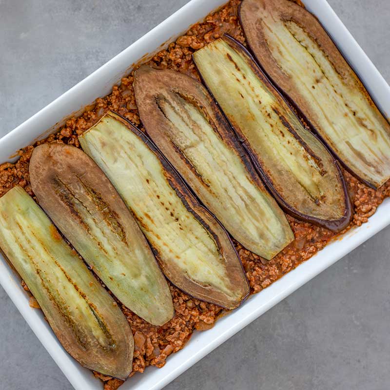 Image of a rectangle casserole dish layered with ground lamb sauce and grilled eggplant, sitting on a grey background