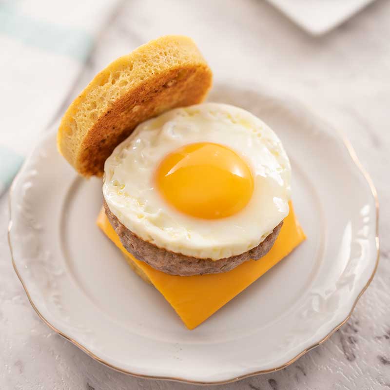 Image shows a keto sausage and egg muffin with the top bread on the side, sitting on a white plate