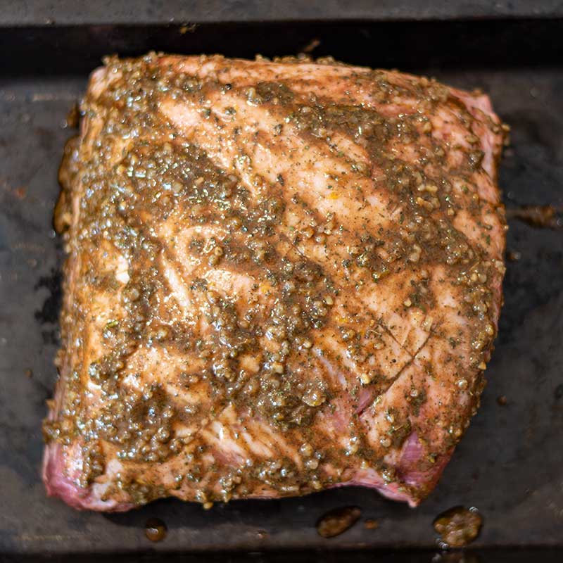 Image show a raw lamb shoulder smothered in marinade in a roasting pan