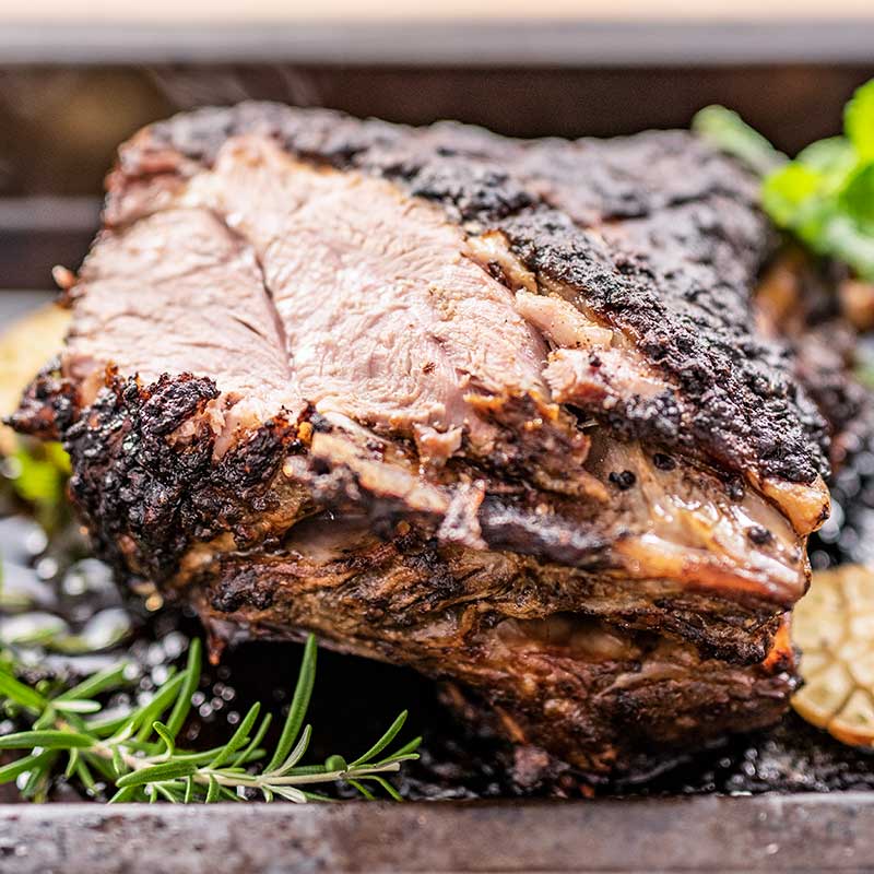 Image show a cooked lamb shoulder in a roasting pan with fresh rosemary, and mint