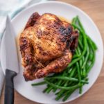 Image of a rotisserie chicken on a white plate with green beans around it on one side and a carving knife on the other.