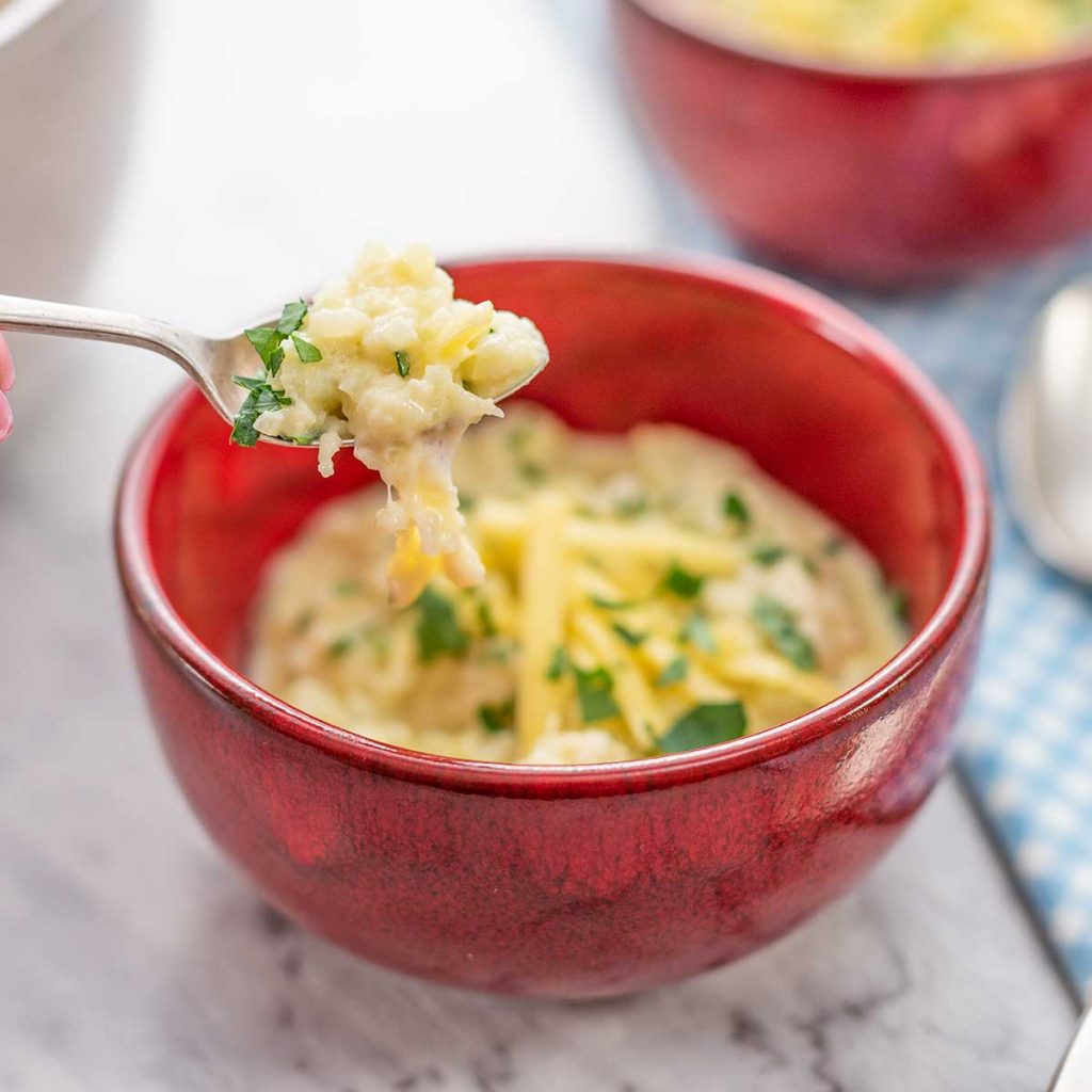 Keto Cheesy Grits in a red bowl, topped with cheddar cheese and chopped parsley, a spoon is lifting out a mouthful