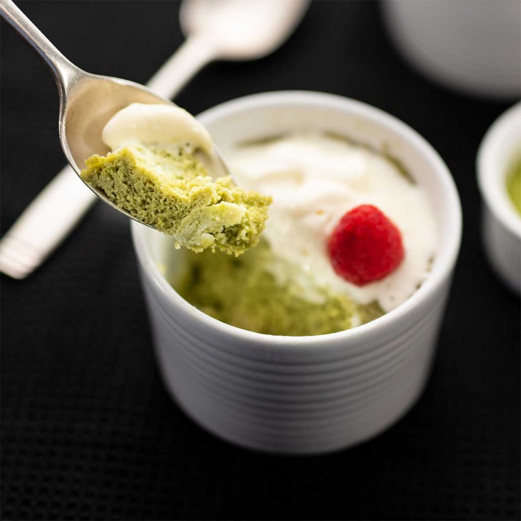 A spoonful of Keto Matcha Mug Cake with whipped cream being lifted from the white ramekin on a black background