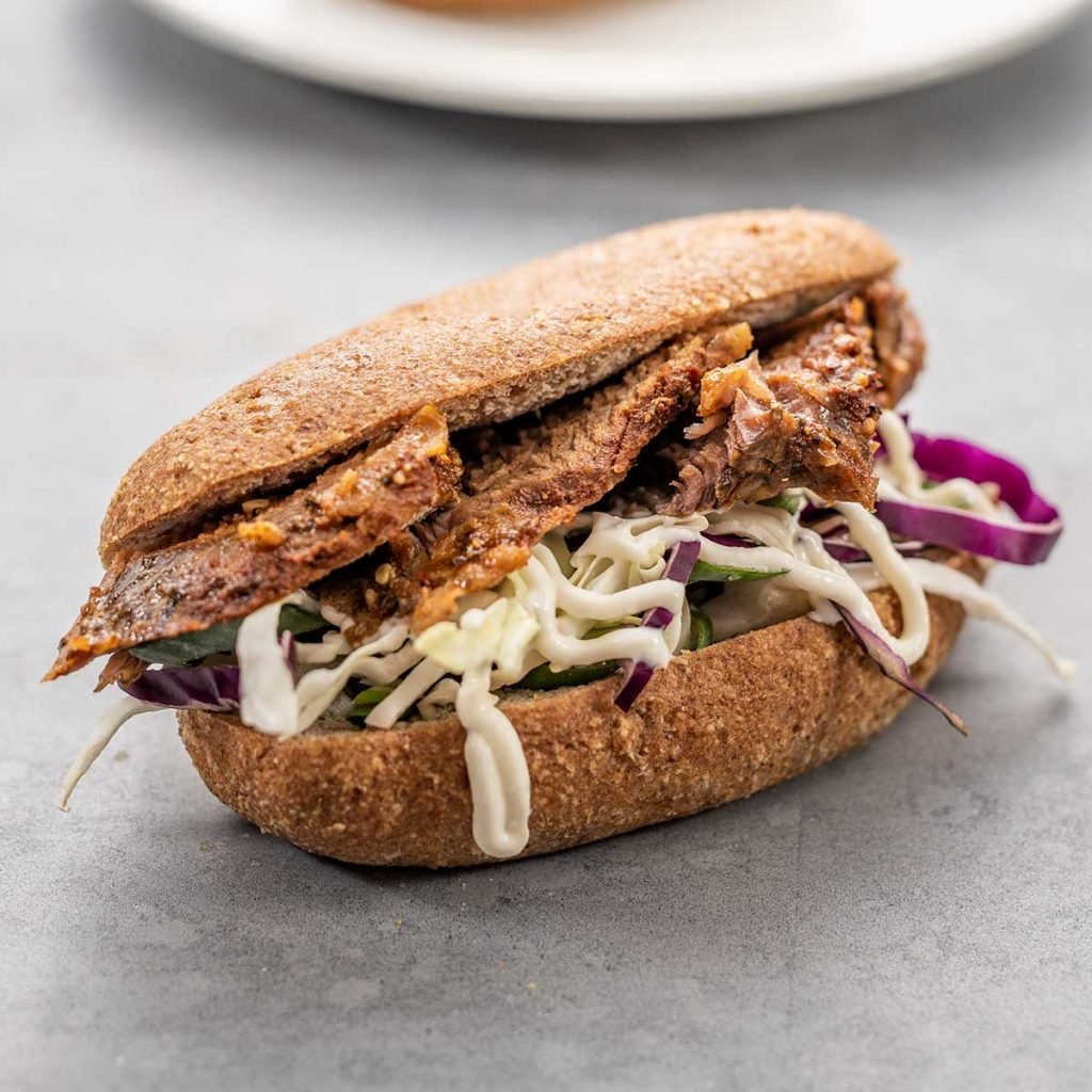 Keto Slow Cooker Tri tip slices on a low car sub roll with coleslaw and mayonnaise on a dark grey surface