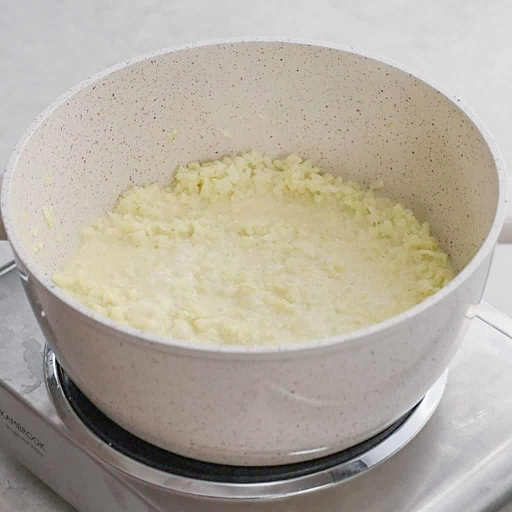 Keto Cheesy Grits cooking on the stove top in a speckled white saucepan