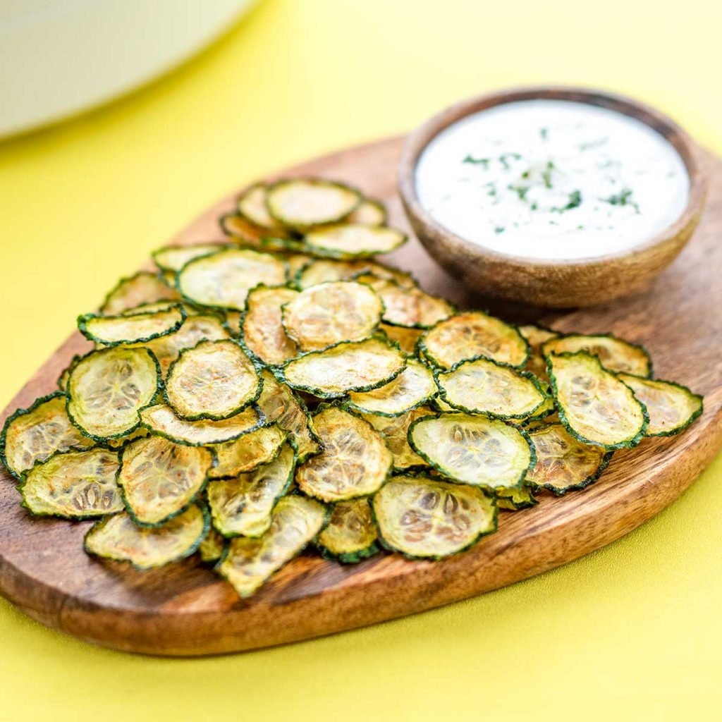 Cucumber Chips on a wooden board with a bowl of creamy dip on a yellow background