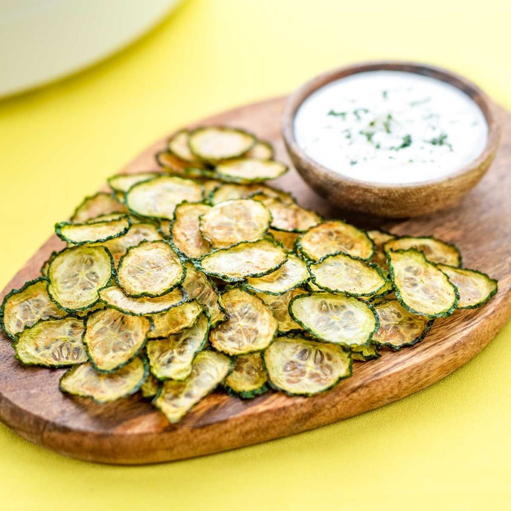 Keto Cucumber Chips on a wooden board with a bowl of creamy dip on a yellow background