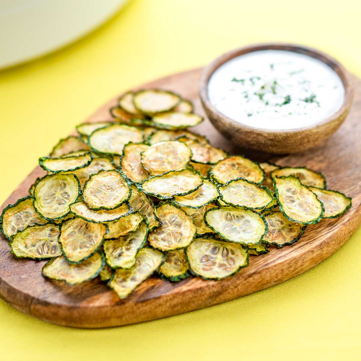 Keto Cucumber Chips on a wooden board with a bowl of creamy dip on a yellow background