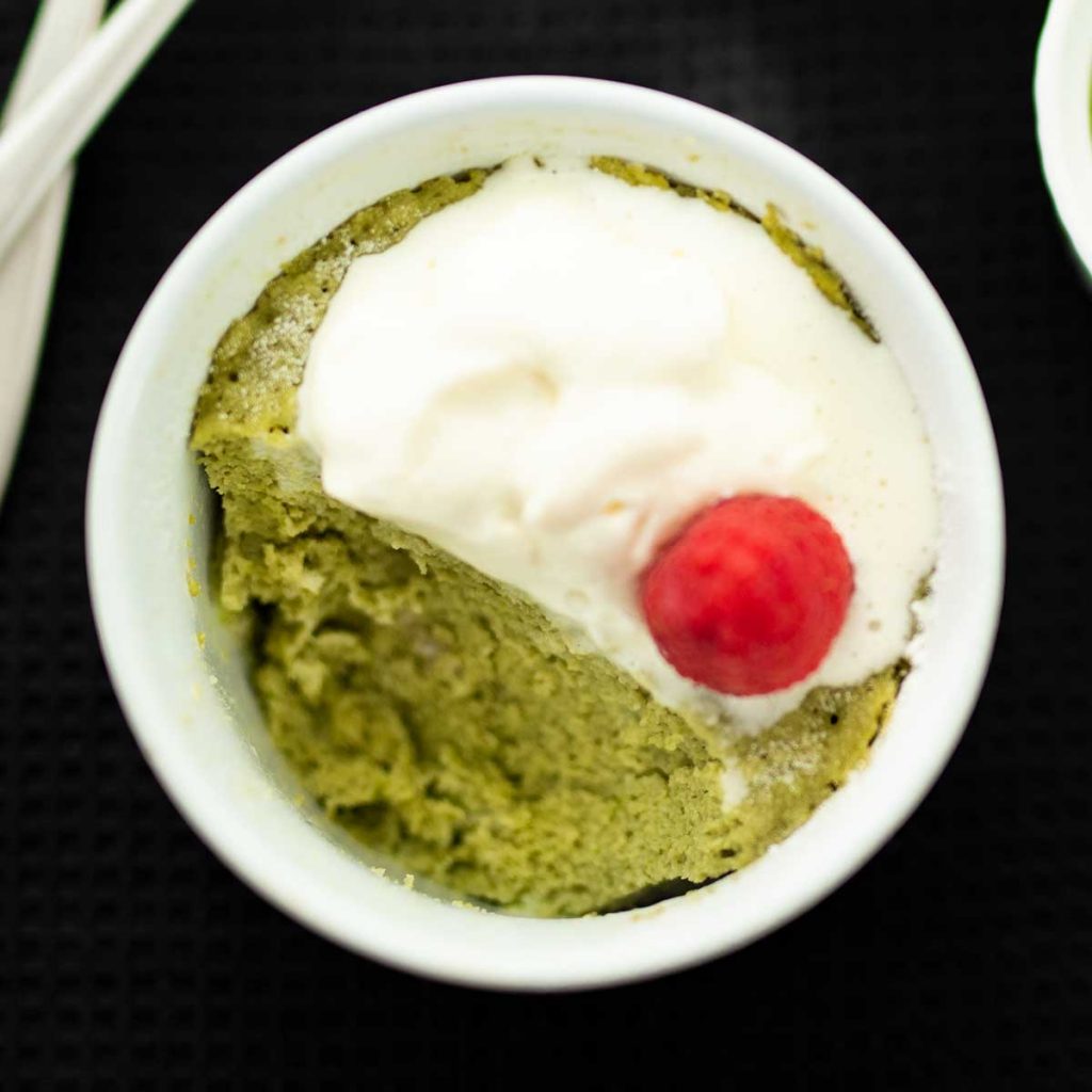 Keto Matcha Mug Cake topped with cream and a raspberry with a spoonful removed in a white ramekin on a black background