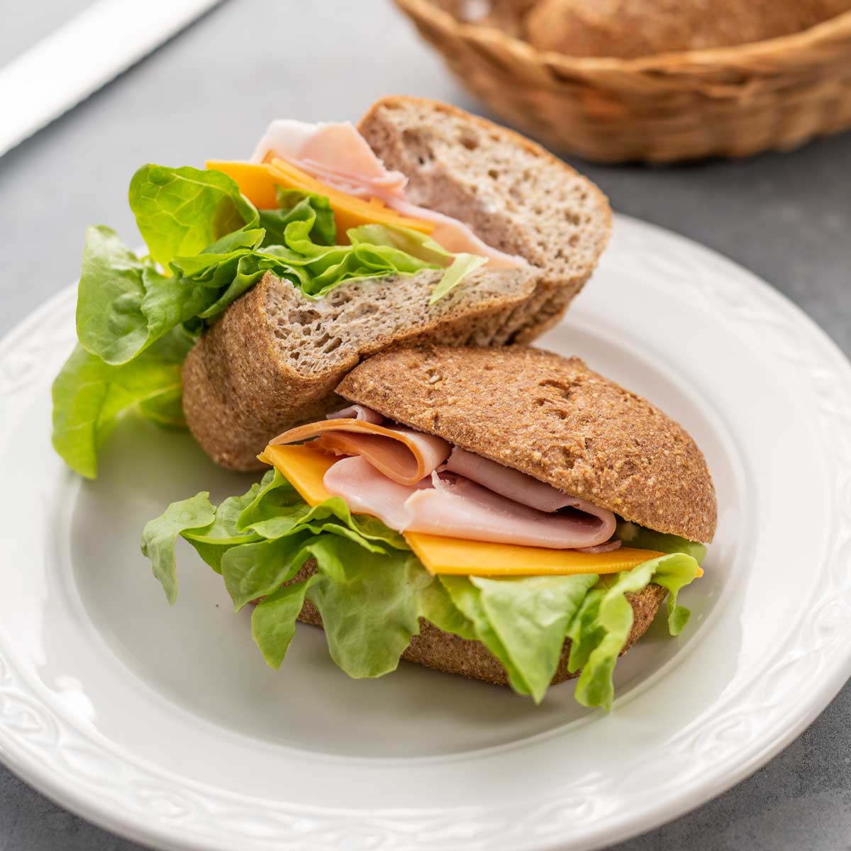 A keto hot dog bun filled with ham, cheddar and lettuce on a white plate