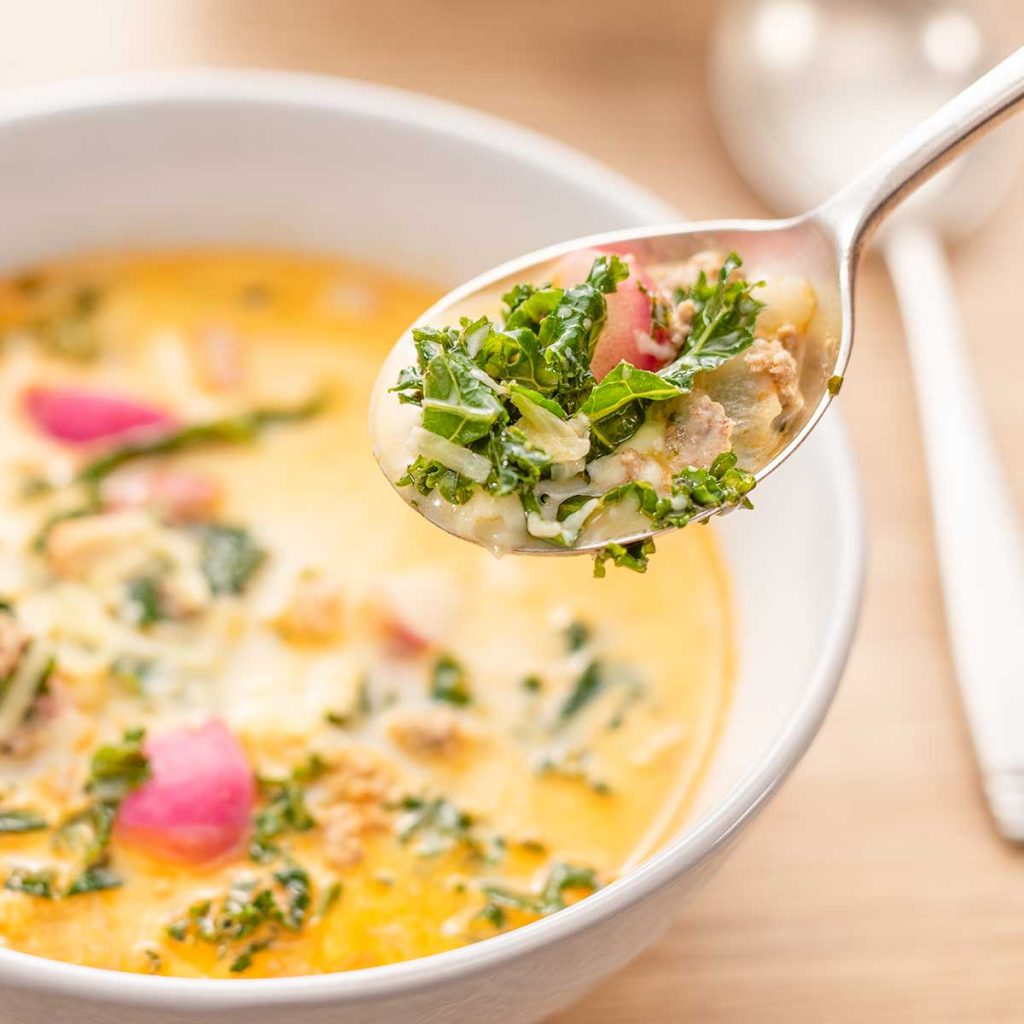 A spoonful of Keto Zuppa Toscana soup being lifted from a white bowl on a wooden table