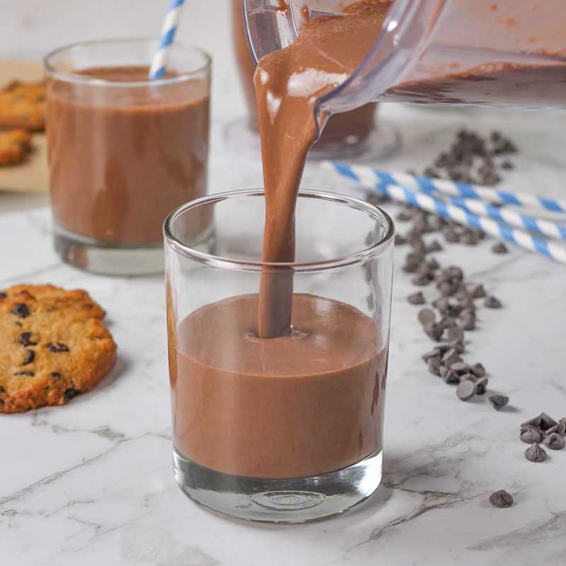  Keto Chocolate Milk being poured into a glass with cookies and chocolate chips in the background
