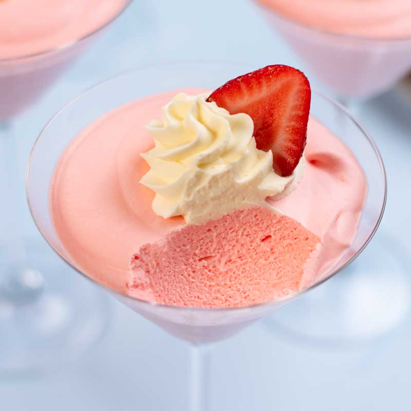 Keto Strawberry Whipped Jello in a martini glass topped with whipped cream and a slice of strawberry with a spoonful removed to show the fluffy insides.