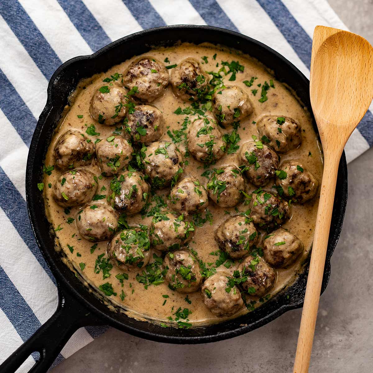 Keto Swedish meatballs in a cast iron skillet on a blue stipe tea towel and a wooden spoon.