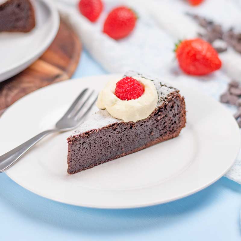 A sliced of Keto Flourless Cake on a plate topped with whipped cream and a raspberry.