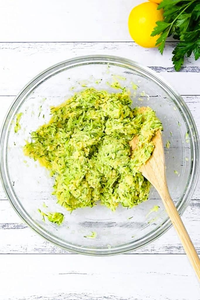 Keto zucchini fritters ingredients in a clear bowl with a wooden mixing spoon.